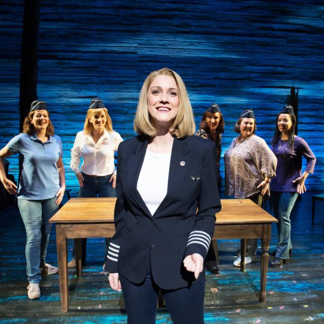 The cast of Come From Away singing Me and The Sky. The female lead is wearing a captain's outfit, and the rest of the cast are standing around a table.
