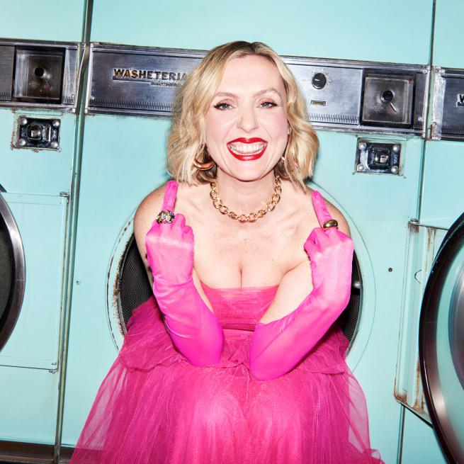 Eleanor in a bright pink dressing, giving the middle finger in a laundrette 
