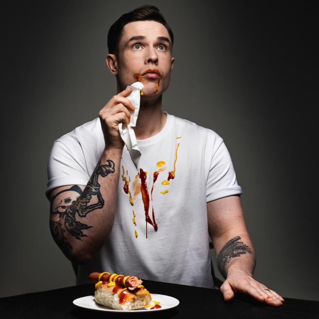 Ed Gamble is wearing a white shirt that is covered with ketchup and mustard while eating a hot dog.