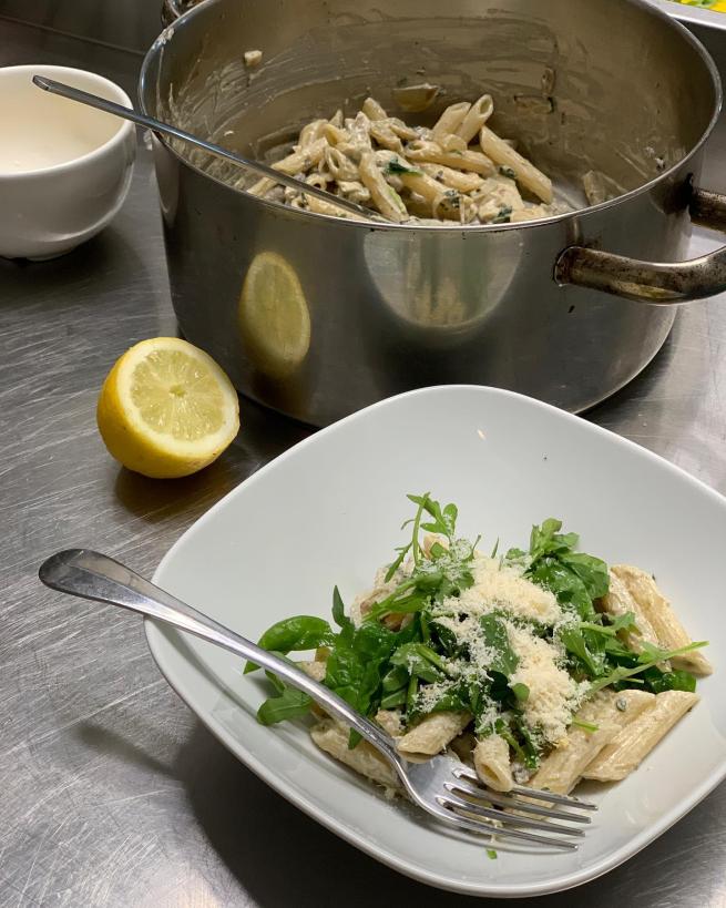 A large cooking pot and a bowl of creamy, cheesy pasta with rocket leaves.