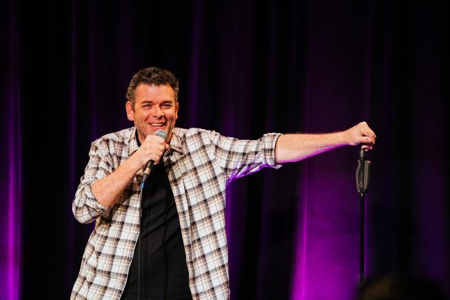 A man wearing a check shirt holding a microphone laughing