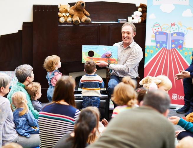 A Leeds Libraries Story & Rhyme session, with a male member of staff reading a book to a large group of children, parents and carers.