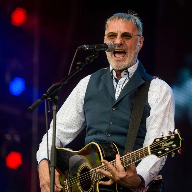 Steve Harley playing a guitar and singing into a mic on stage. 