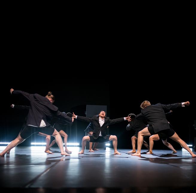 A group of dancers on stage wearing suits with lights shining forward and one dancer screaming in the centre