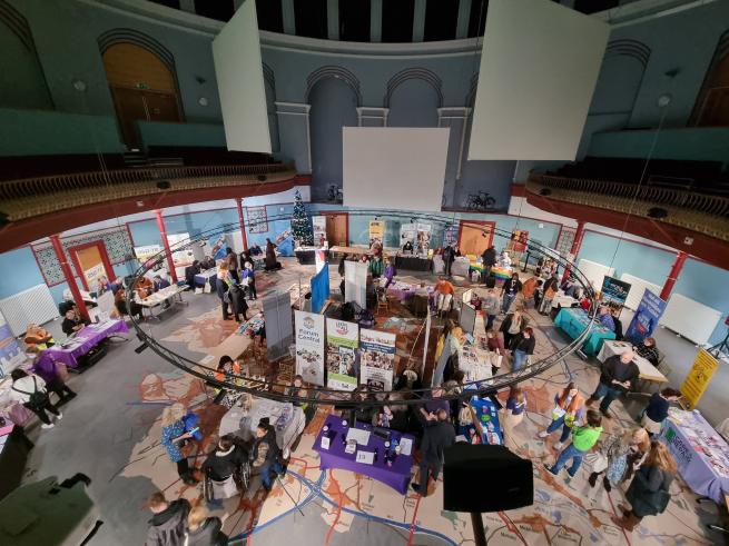 Photo of the 2022 IDODP event from high up in Leeds City Museum, looking down at the event taking place below. In the large hall there are stall all around the room with lots of people talking to one another at the stalls and mingling. It seems like a hive of activity is taking place.
