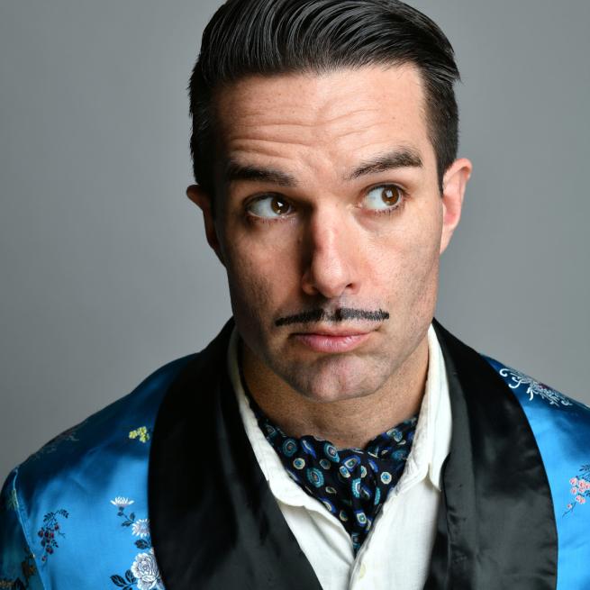Troy Hawke wearing a blue robe and a drawn moustache. 