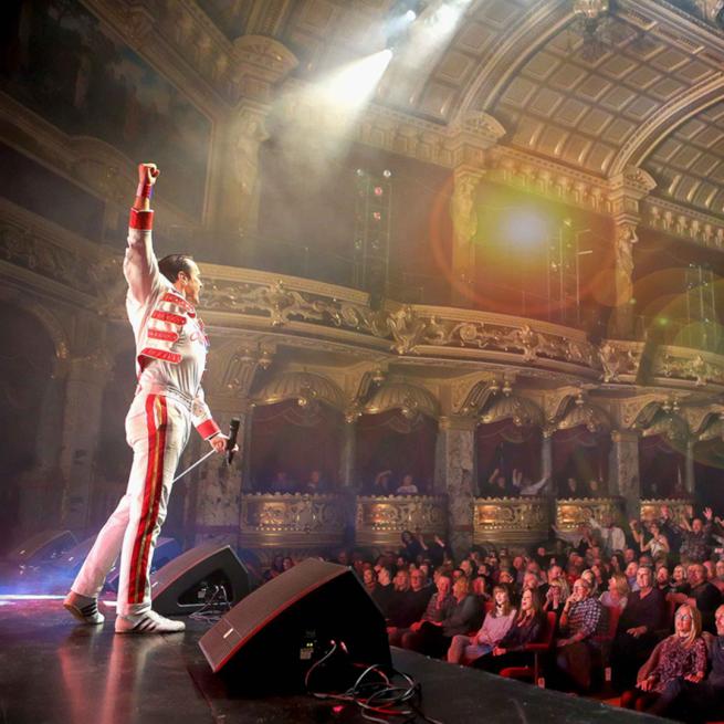 A performer on stage dressed as Freddie Mercury. He is performing to a full crowd. 