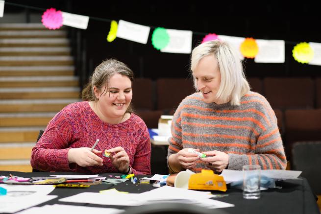 two people are in the middle of a craft project, sat in front of a desk, smiling at each other.