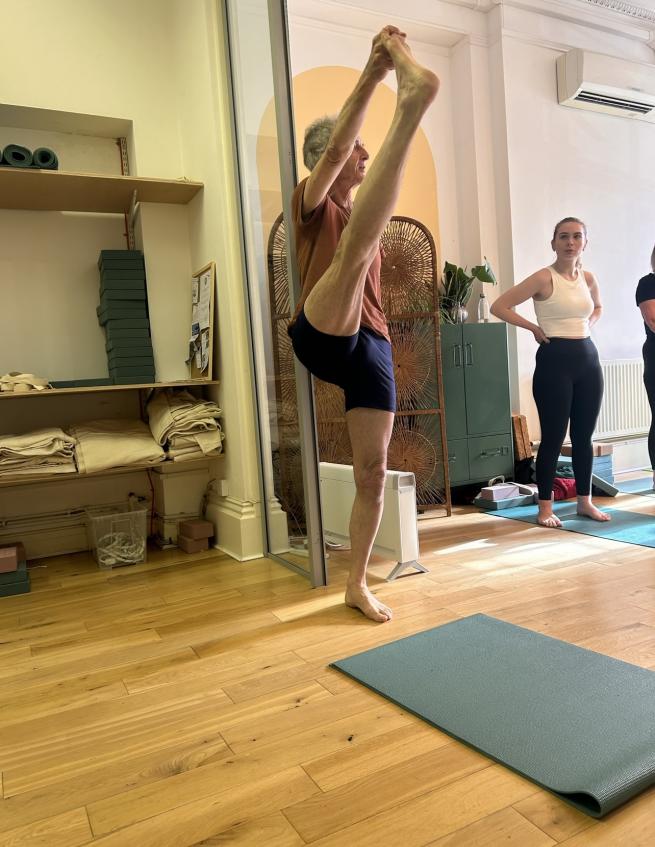 Alan is doing a yoga pose lifting one leg and holding his toe with straight legs whilst students look