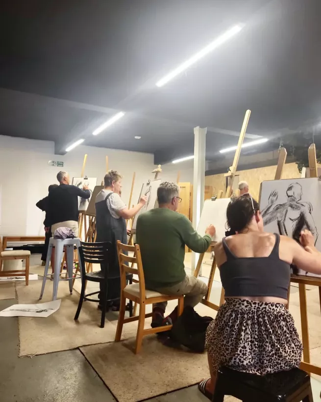 An art studio room of people sat at easels using charcoal to draw a life model 
