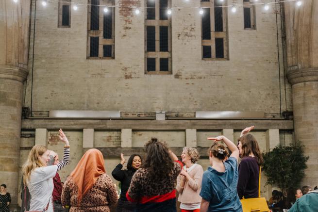 A diverse group of women dancing in a circle, some have their arms raised, they are laughing 