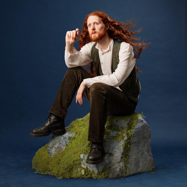 Alasdair sits on a moss-covered rock. He is wearing a vest and corduroy trousers. He has long wavy red hair and a red beard.