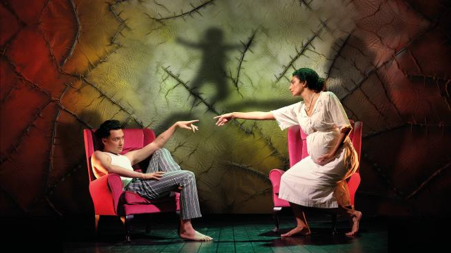 A man and a woman sit in red arm chairs. They reach towards each other extending their index fingers.
