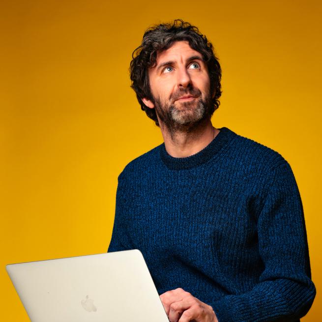 Mark Watson is sitting and working on his apple computer. He is looking off into the corner. He is wearing a blue jumper.