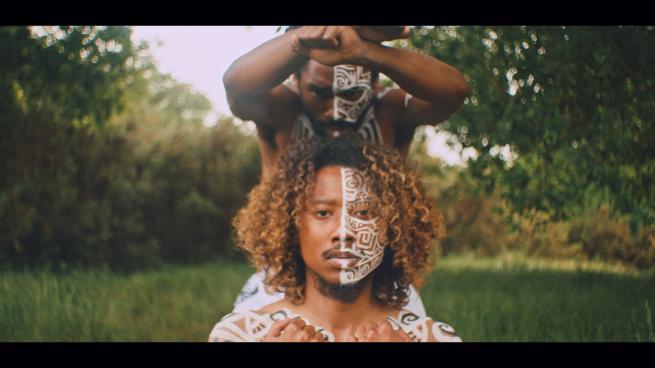 Exterior, background shows green field and trees. Daytime. Two men stand front and centre of the image, one immediately behind the other. Their faces serious. Both are topless, their upper torso and faces painted with white patterns. 