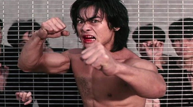 Man with a bloody mouth and fists raised in front of a fence behind which are more men.