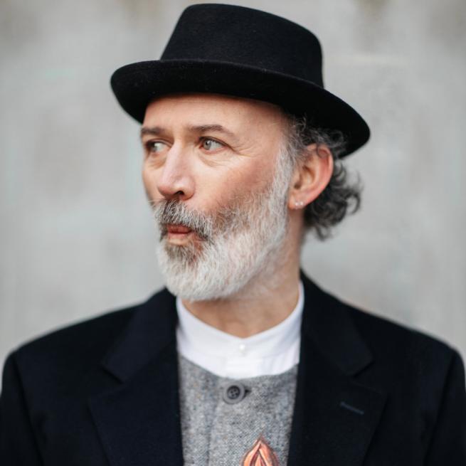 Tommy Tiernan is wearing a black fedora and jacket. He has a white beard and is pursing his lips.