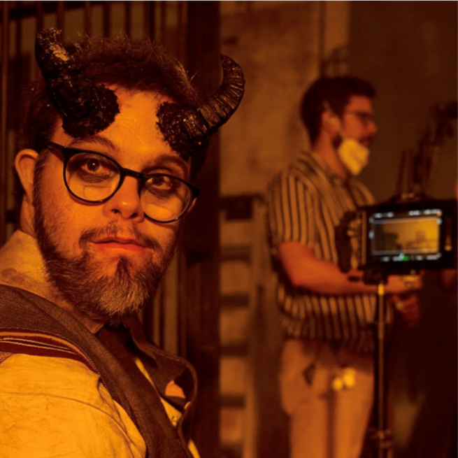 Otto Baxter looks over his shoulder pat the camera with a pair of devil-style horns appearing to grow from his head. In the background is a camera viewfinder and a crew member of a film crew talking to someone out of shot.