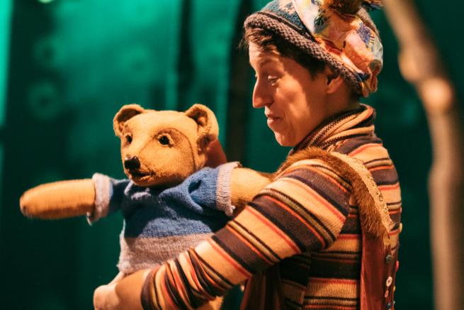 A person wearing a stipey top holds a teddy bear wearing a blue jumper out to face the audience