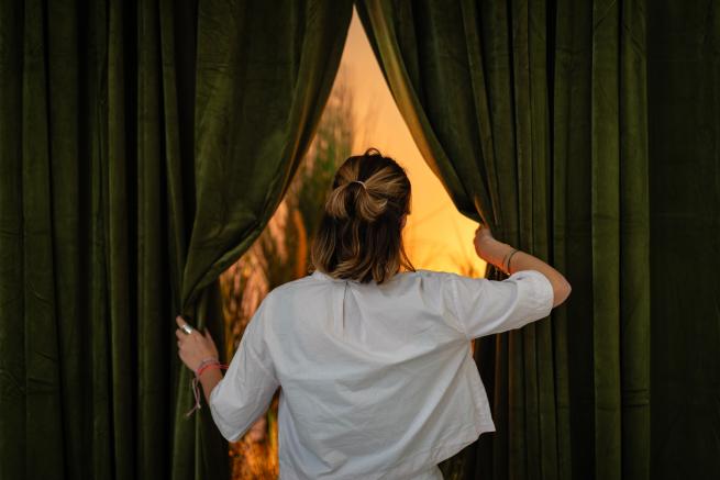 A young woman in a spa, linen shirt reaching through a curtain which has light coming from behind it. 