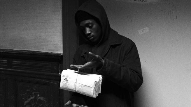 black and white, a black man in a hooded top holds a bundle of letters.