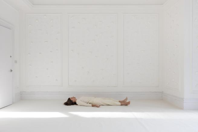 Artist Gemma Anderson-Tempini lying on a white carpeted floor in a room featuring white wallpaper of geometric polytopes, some of which protrudes from the flat surface into the third dimension.