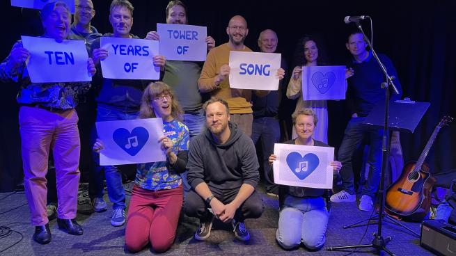 Eleven Tower Of Song performers standing on stage holding sheets of paper with the words "Ten Years Of Tower Of Song" on them.