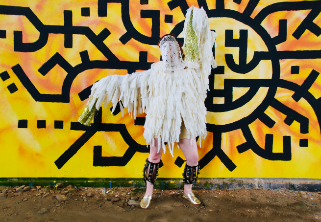 A photograph of a female dancer wearing a white ragged costume, standing in front of a yellow and black graffitied mural