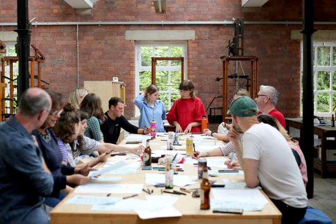 Eleanor and Georgia stand at the head of a long table lined with workshop participants. The table if filled with paper and drawing materials.