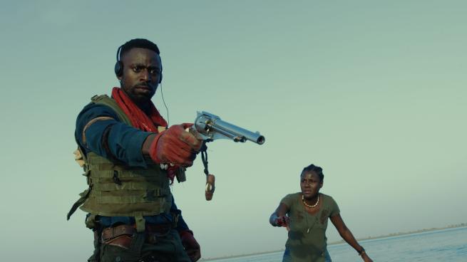 Man in military outfit pointing gun at something with woman in the background watching, in front of a body of water.
