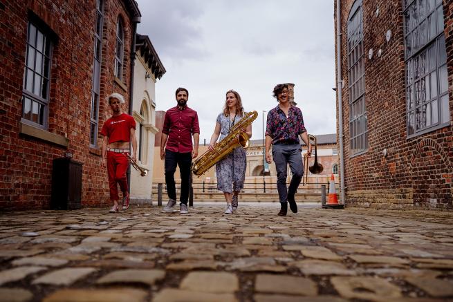 4 musicians walk down a cobbled street holding their instruments.