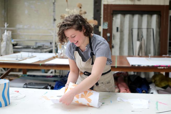 Millie is stood wearing a well used apron as she cuts away fabric from her screen printed sample. She has printed onto white linen with peach coloured flowers.