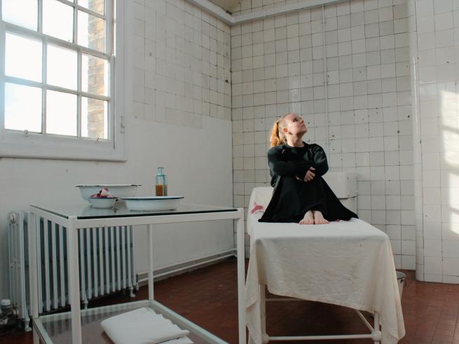 Playwright and musician Amanda Grace sitting on a bed in a tiled Victorian hospital.