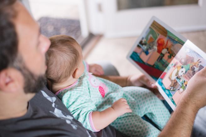A man with light skin, brown hair, and a short beard is holding a baby on his lap. The baby is wearing a pale blue sleep suit. Both of their faces are in profile and they are both looking at a picture book with colourful images of animals.