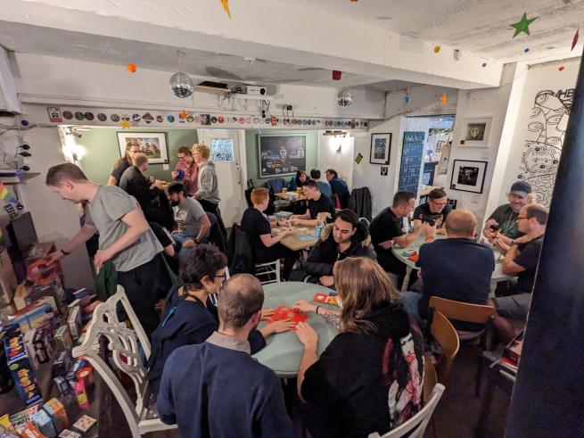 A room full of people - most are sitting at tables playing games - some are standing up either choosing games or going to the bar.