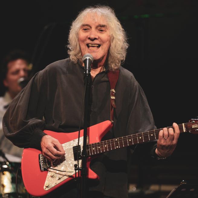 Albert Lee on stage singing into a microphone and playing a red electric guitar.