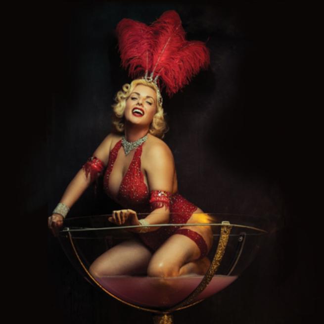Burlesque dancer in a red leotard and a red feather head piece sat in a giant martini glass.