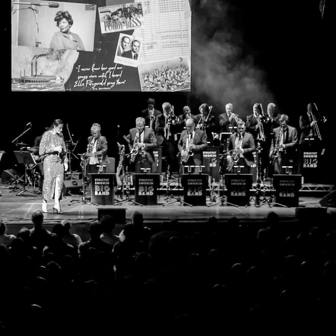 A black and white photo of a woman in a sparkly dress on stage with the Strictly Smokin Big Band in front of an audience. Behind the band is a projection of images including Ella Fitzgerald, Porgy and Bess and a factory.
