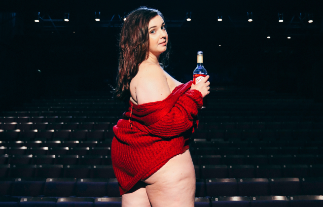 A person on stage wearing a red jumper and holding a bottle. In the background, the auditorium of a theatre.