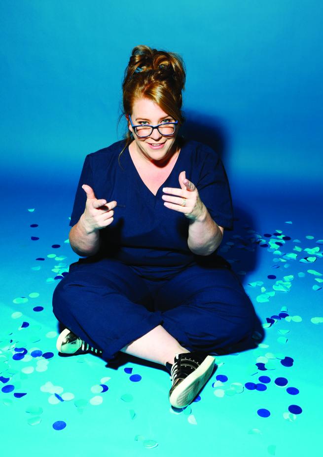 Georgie Carroll in dark blue scrubs sat cross-legged on the floor pointing at the camera with both hands and surrounded by big circles of blue confetti. 