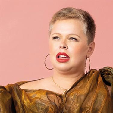 Laura Smyth in a brown dress and hoop earrings looking off to the side in front of a pink background.