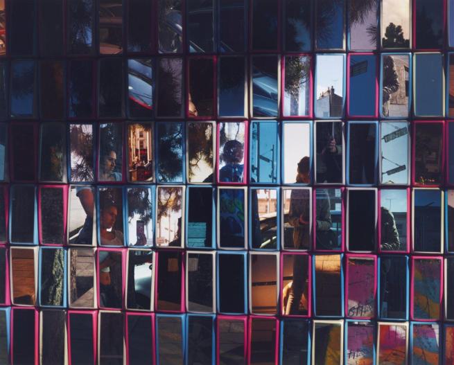 A colourful fragmented photograph. The artist Hannah Starkey captures herself in the act of taking a photograph. Her reflection, as well as those of two young women and the anonymous street they stand in, is presented to the viewer fractured and distorted in the mirror’s multiple planes.