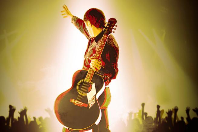Man with a guitar faces away from the camera towards silhouettes of hands and arms in the air to indicate an audience. 