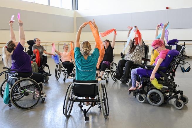 A group of wheelchair users in a circle, raising their arms with brightly coloured scarves