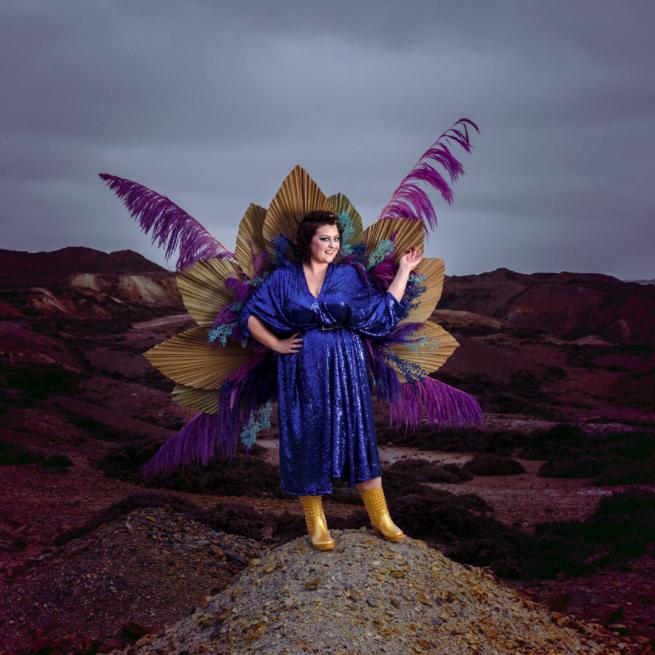 Kiri Pritchard-Mclean standing on a rock in a moody outdoor landscape. She wears a blue dress and gold wellies with large purple and gold feathers all around her.