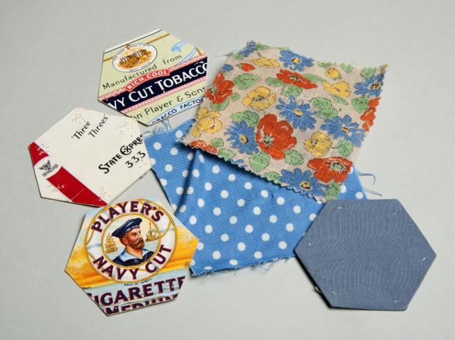 A photograph of card hexagons made from Navy Cut cigarette packets, and pieces of blue, polka dot and floral patterned cloth, used to make patchwork in the Second World War.