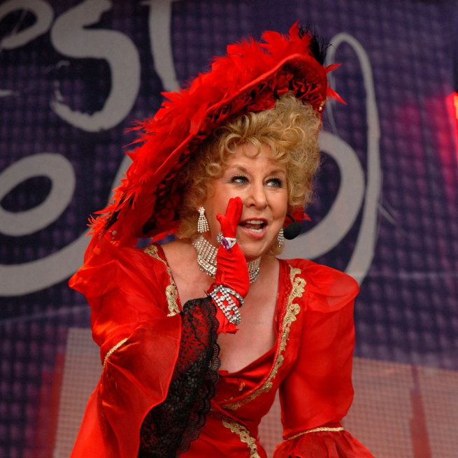 Jan Hunt wearing a very big red hat and red dress looking like she's whispering with her hand at her mouth with a stage screen in the background.