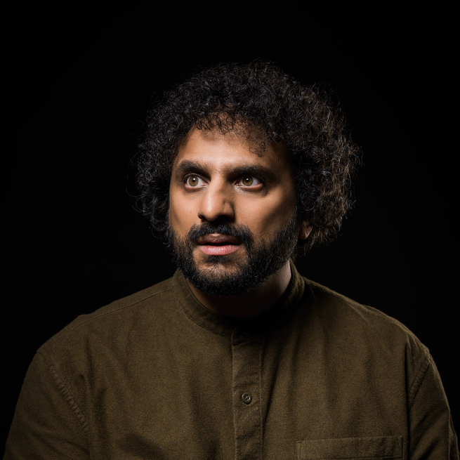 Nish Kumar in an olive buttoned up shirt with a black background.