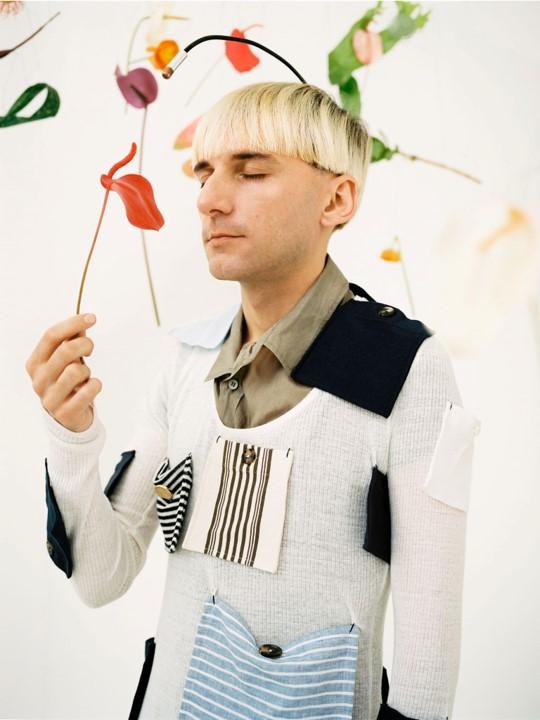 Neil Harbisson stands with eyes closed holding a red flower up to the antenna which is implanted in his skull