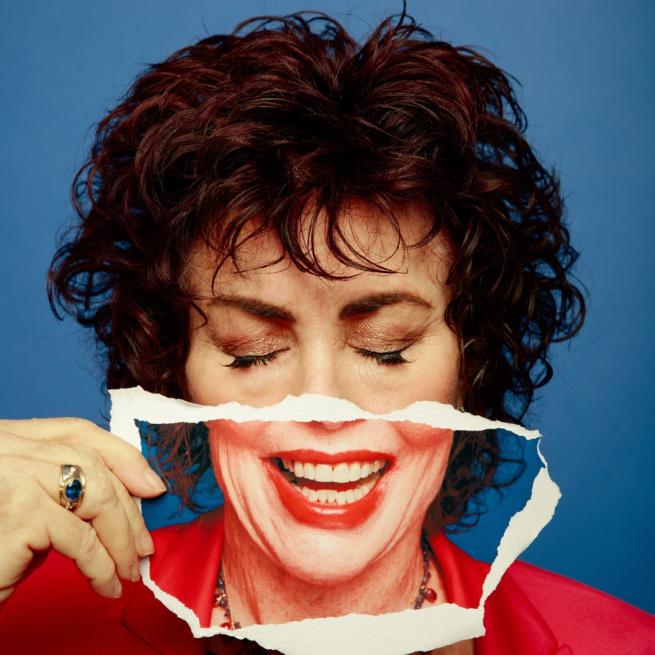 Ruby Wax with her eyes closed holding up a torn photo of a smiling mouth to the lower half of her face.